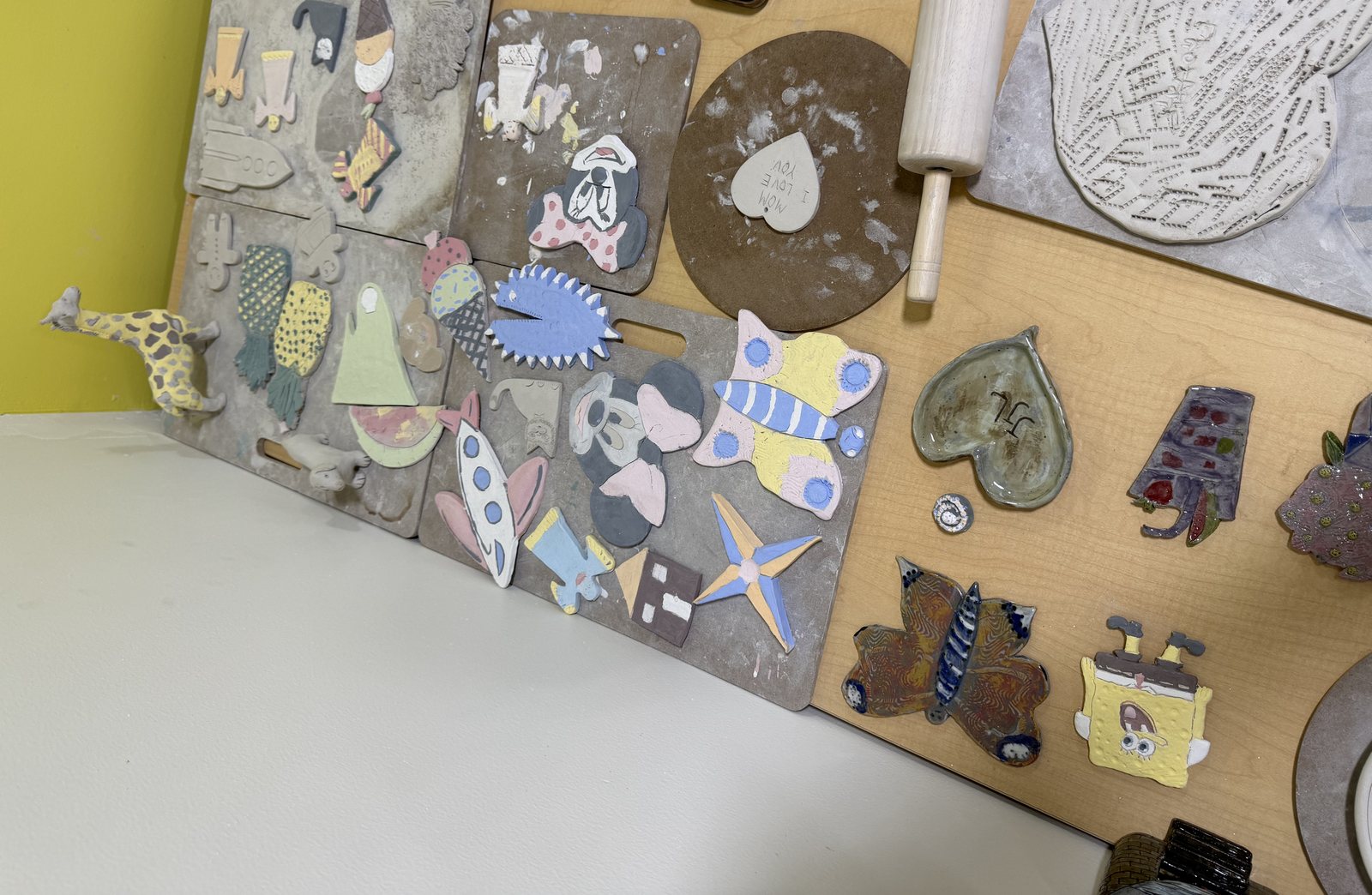 A studio table covered with hand-made clay pieces at different stages — a giraffe, a rocket, butterflies, a gnome, a heart dish and a hand-lettered “Mom I love you” tile, some still drying and some painted